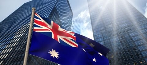 Two glass skyscrapers reaching a clear blue sky. In front of them, the Australian flag.